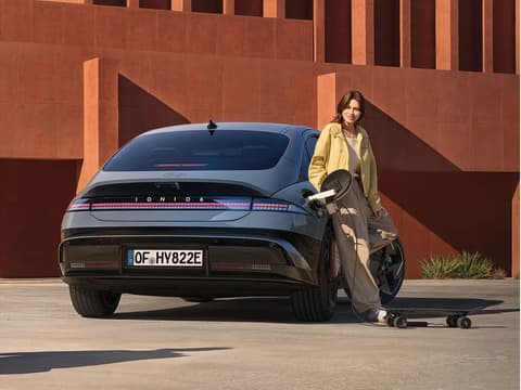 Жена, опряна на IONIQ 6, зарежда електрически скейтборд, включен във външния заряден порт.