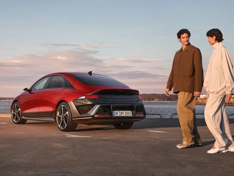 Мъж и жена минават покрай червен Hyundai IONIQ 6 N Line, паркиран до океана.