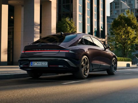 HYUNDAI IONIQ 6 преминава по модерна градска улица, заснет отзад със стилен аеродинамичен дизайн и градска архитектура на заден план.