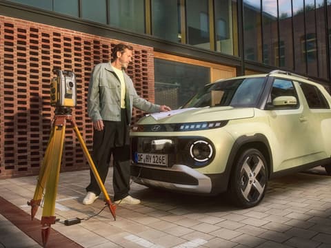 Външен порт Vehicle-to-Load на Hyundai INSTER, който захранва геодезично оборудване; до него стои мъж.