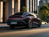 HYUNDAI IONIQ 6 преминава по модерна градска улица, заснет отзад със стилен аеродинамичен дизайн и градска архитектура на заден план.