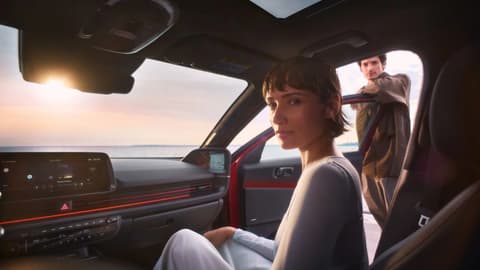 Жена на предната пътническа седалка на Hyundai IONIQ 6 с мъж, който се опира на отворената врата.