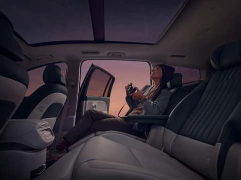 Човек с камера на задната седалка на Hyundai IONIQ 9, вратата е отворена към лилаво-розово небе.