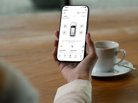 Ръка държи смартфон с приложение за управление на електромобил – с дистанционни настройки на климатика и състояние на автомобила.