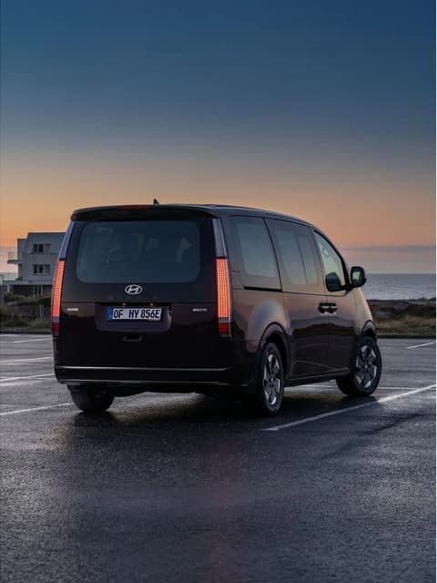 Гледка отзад на Hyundai STARIA Electric, паркиран до модерни къщи край плажа.