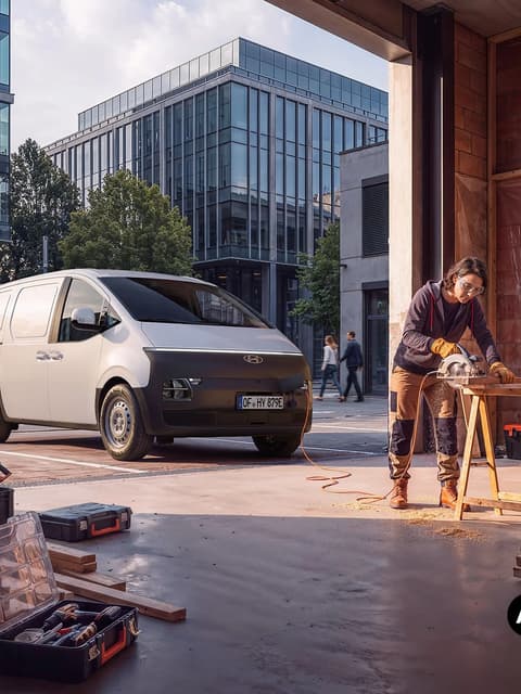 Hyundai STARIA Cargo до работилница, докато работник реже дърво на строителна площадка в града.
