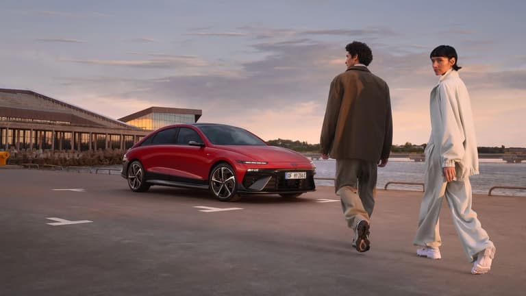 Мъж и жена вървят покрай червен Hyundai IONIQ 6 N Line, паркиран до океана.