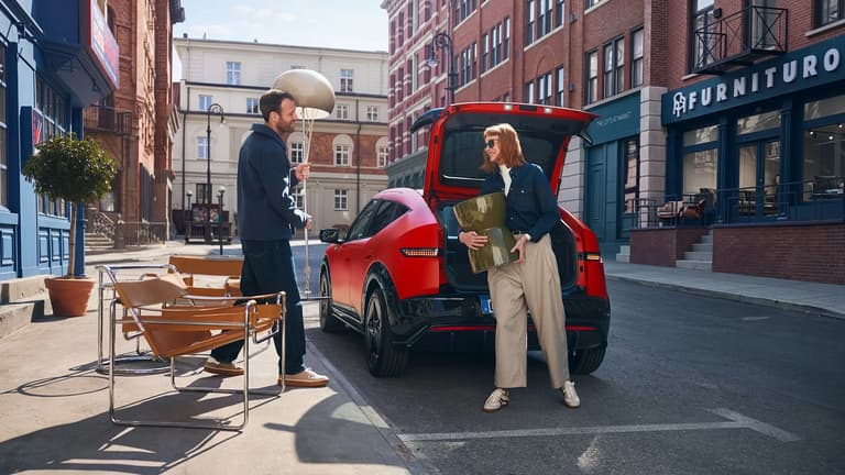  Two people unload furniture from the trunk of a red Hyundai IONIQ 3 on a city street..