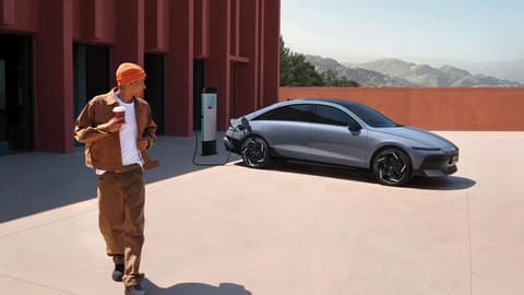Hyundai IONIQ 6 се зарежда на станция до модерна сграда, докато човек преминава наоколо