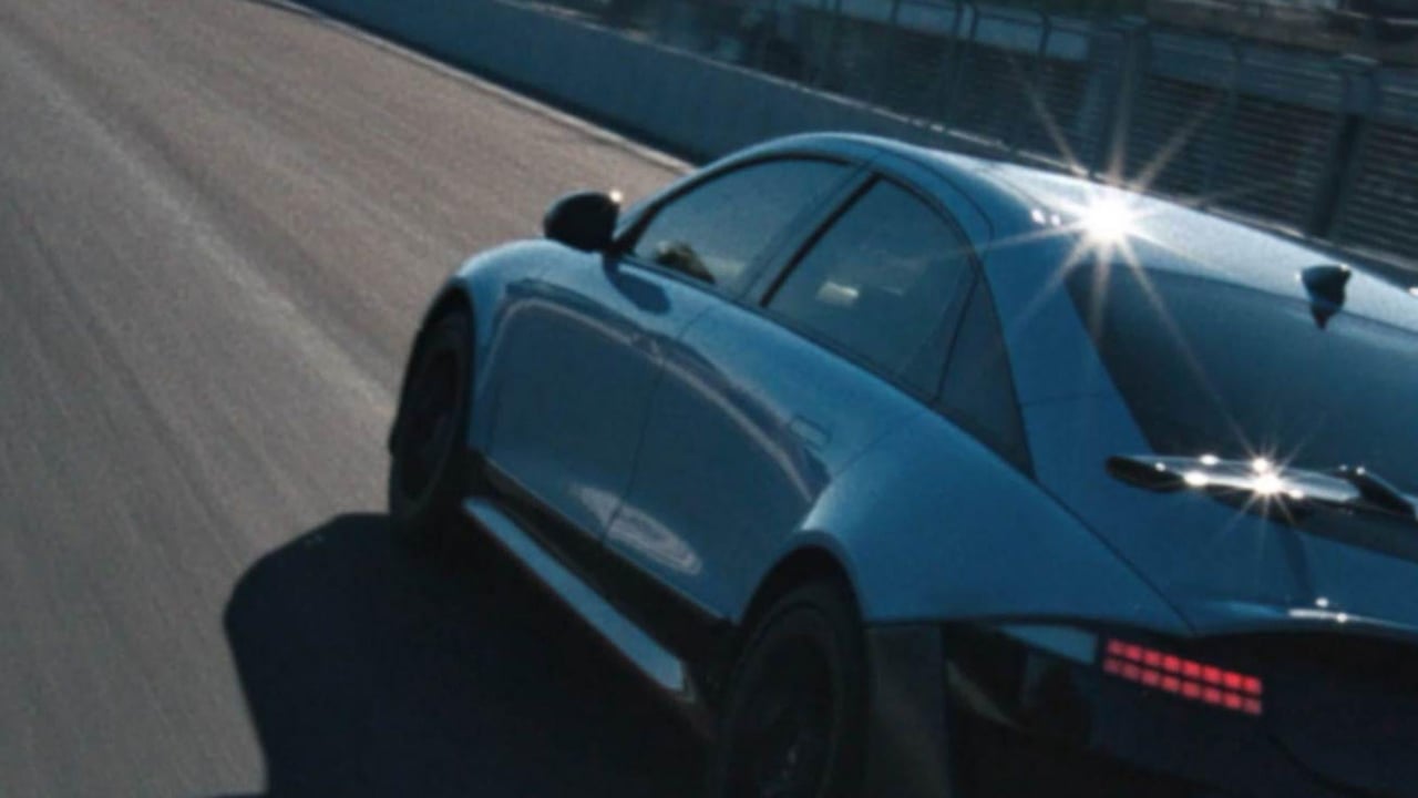 A Hyundai IONIQ 6 N speeds past on the track, reflecting sunlight with a modern building in the background.
