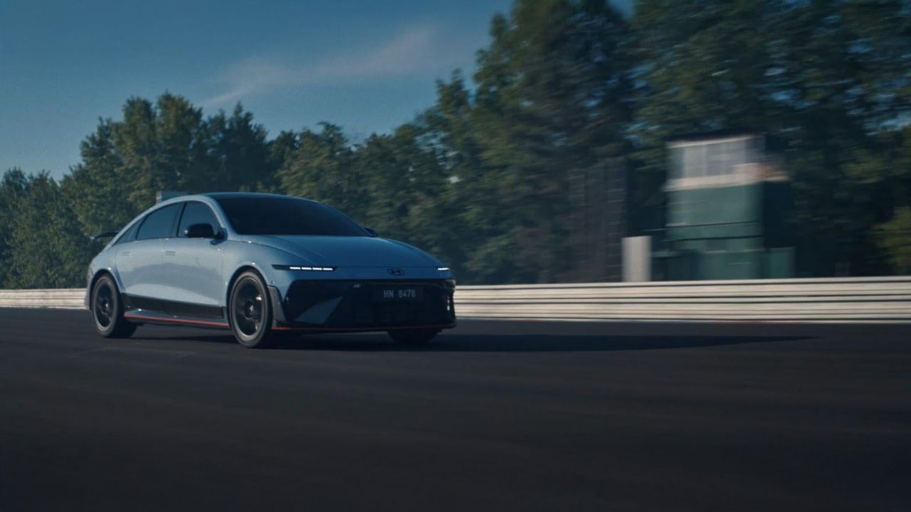 A Hyundai IONIQ 6 N electric performance car speeds along a racetrack with trees and a building in the background.
